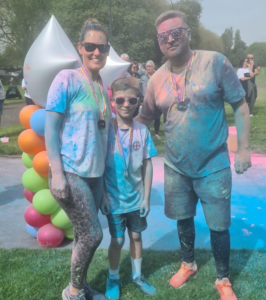 This shows three people, a man, woman and young child at a colour run in aid of Beacon. They are covered in brightly covered paint powder.